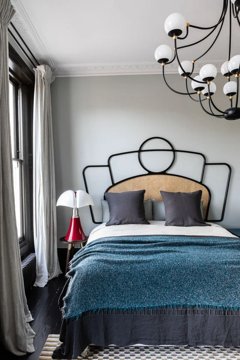 A bedroom with a bed that has a black metal headboard with geometric shapes, two brown pillows, and a teal throw blanket. To the left, there's a small bedside table with a red and white lamp. The room has large windows with cream-colored curtains and a modern black chandelier with white globes hanging from the ceiling.