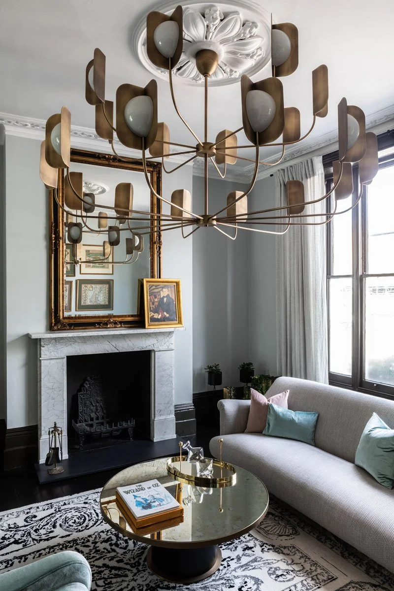 Living room with a modern chandelier, a fireplace with a gilded mirror above it, a beige sofa with pastel pillows, a black and white patterned rug, a round coffee table with magazines, and a large window with curtains.