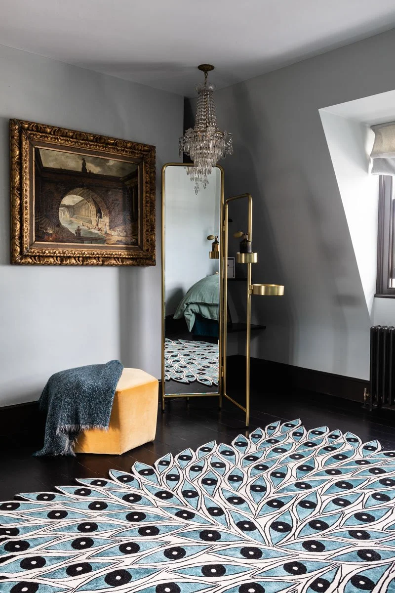 Interior view of a stylish room featuring a large, decorative area rug with a peacock feather pattern, a gold-framed standing mirror, a crystal chandelier, a framed painting on the wall, a small yellow ottoman with a gray throw blanket, and a black side table with yellow lamps. There is a window with a white Roman shade and a black radiator beneath it.