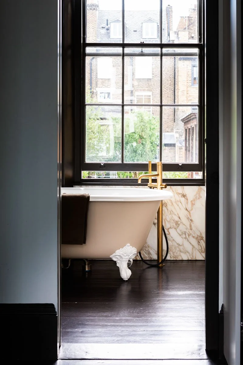 A bathroom with a clawfoot bathtub positioned in front of a large window with multiple panes, a gold faucet, dark wooden floor, and a marble accent wall.
