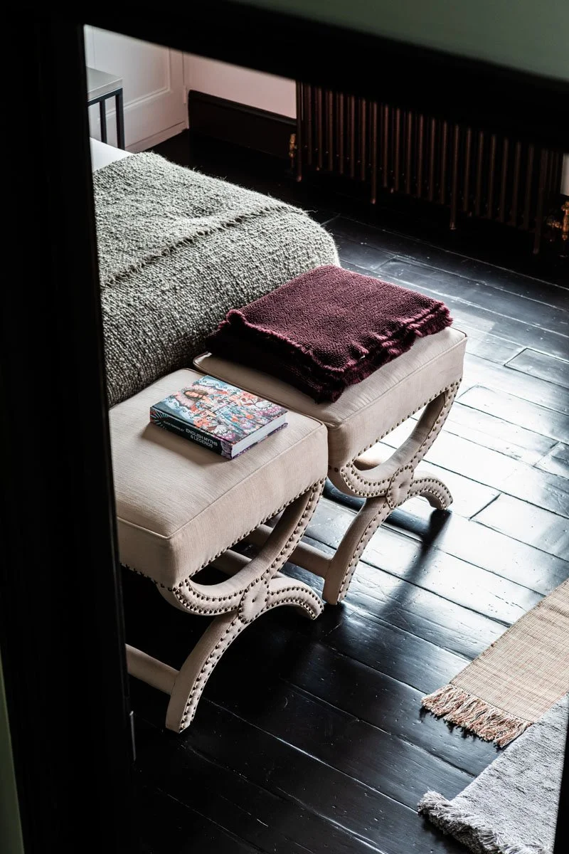 A cozy bedroom corner with a bed, a beige upholstered bench with curved wooden legs, a burgundy throw blanket, a colored book, and a small rug on dark wooden floorboards.