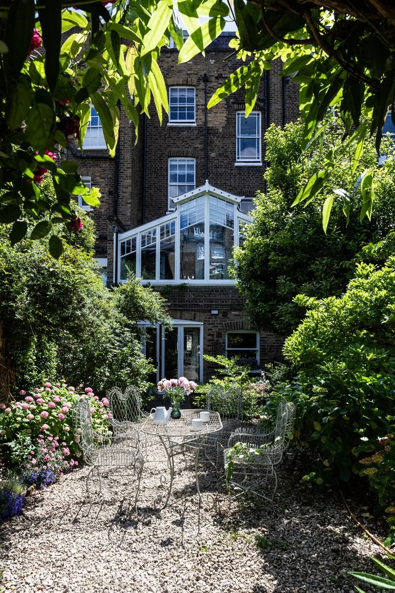 A garden with a round, white metal table and six matching chairs, decorated with a vase of pink flowers and white teacups, surrounded by lush green bushes and flowering plants, with a brick building and glass conservatory in the background.