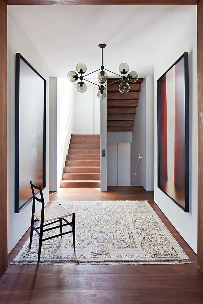 Entryway with wooden floor, modern chandelier, staircase, large abstract paintings, and a chair on a patterned rug.
