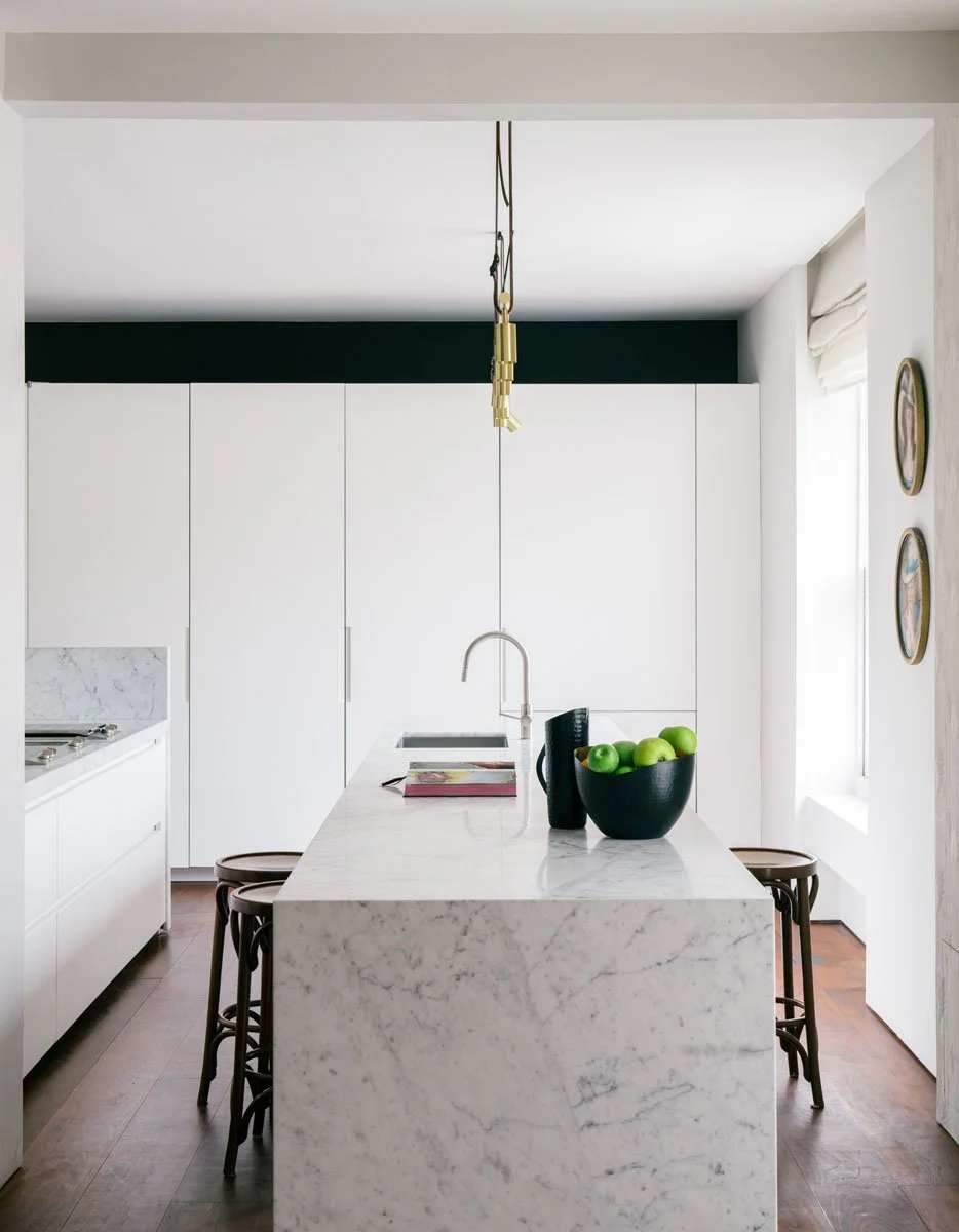 Modern white kitchen with marble island, black bowl of green apples, and wooden stools, illuminated by natural light from the window.
