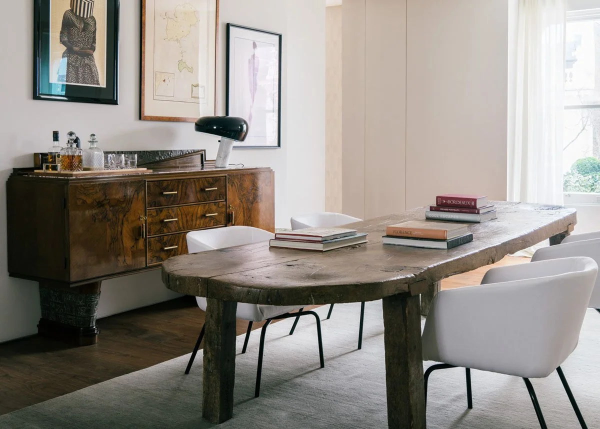 A dining room with a rustic wooden table, four white chairs, a wooden sideboard with bottles and glasses, framed artwork and a map on the wall, and a window with sheer curtains allowing natural light in.