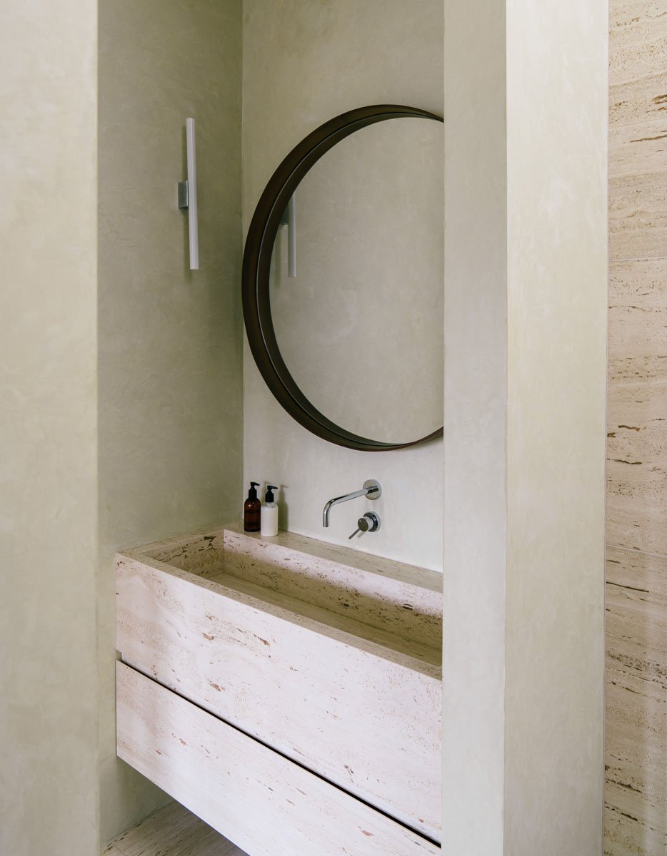 Minimalist bathroom with a rectangular stone sink, wall-mounted faucet, round mirror, and ambient lighting on beige walls.