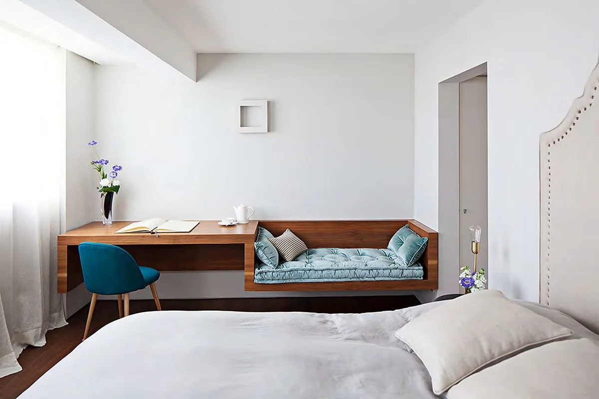 Minimalist bedroom with a white bed in the foreground, a window with sheer curtains, a wooden desk with a blue chair, a vase with purple flowers, an open book, and a white teapot on the desk. A built-in seating area with blue cushions is on the wall, next to a small lamp and flowers.