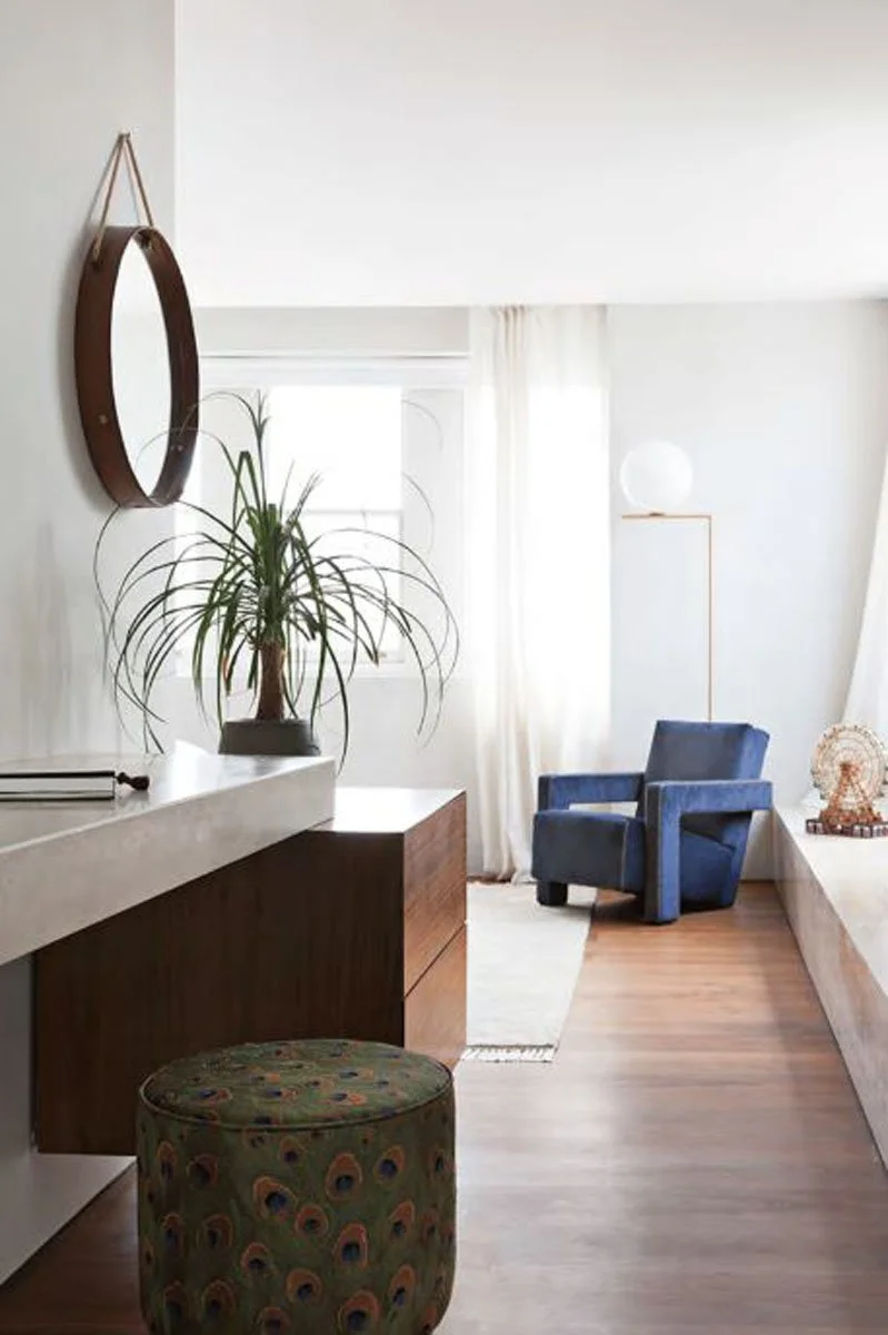 Interior view of a bright living room with a blue armchair, white curtains, a potted plant, a mirror on the wall, and decorative items.