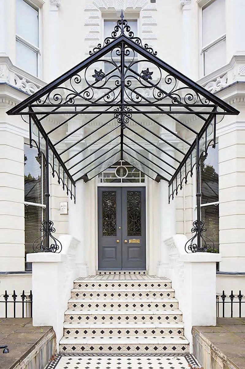 Front entrance of a white building with black decorative ironwork and a dark blue door, accessed by a short flight of tiled stairs.
