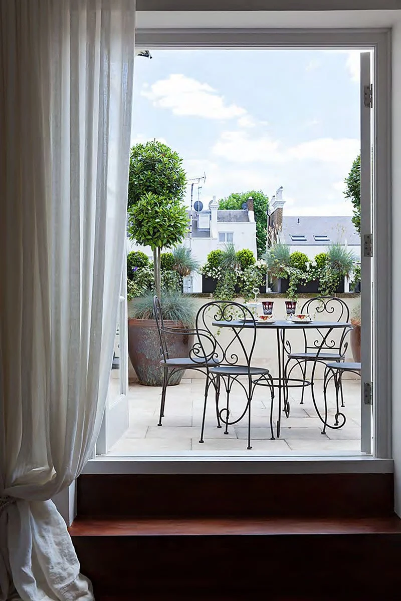 Open doorway leading to a patio with a round metal table and three matching chairs, surrounded by potted plants and a low wall with flower boxes, under a partly cloudy sky.