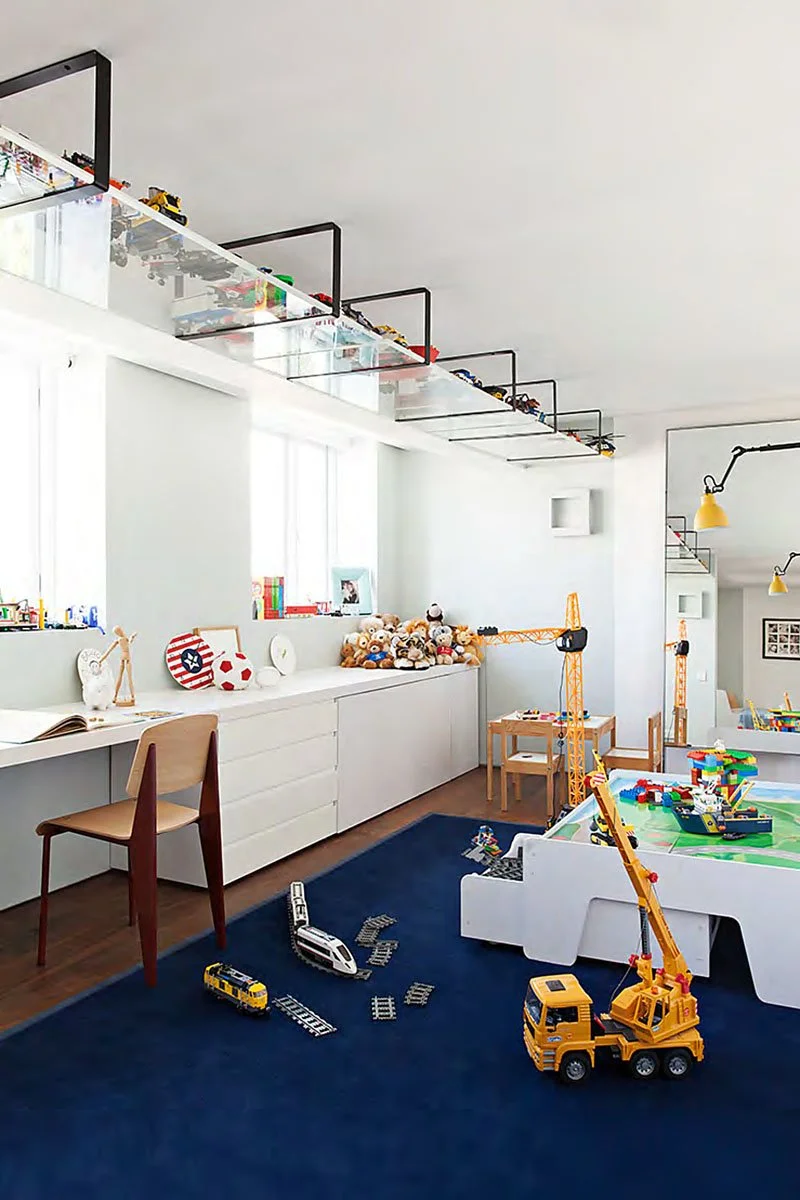 A playroom with a blue rug, various toy vehicles, teddy bears, LEGO blocks, a small desk, and toy construction cranes, with toy cars and trucks on a white table and shelf. The room has white walls and large windows, with yellow pendant lights hanging from the ceiling.