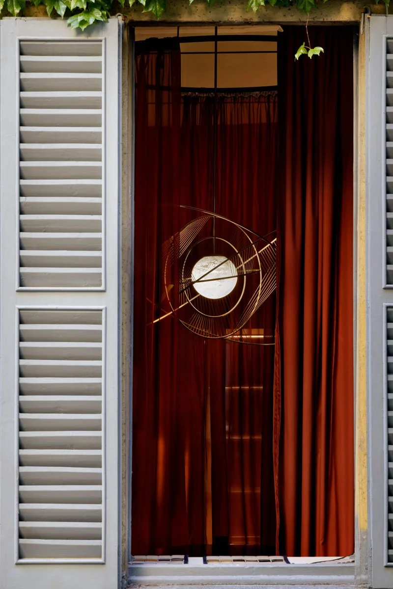 A view through a window with cream-colored shutters, showing red curtains inside and a modern, artistic light fixture with a geometric design and a circular shape.