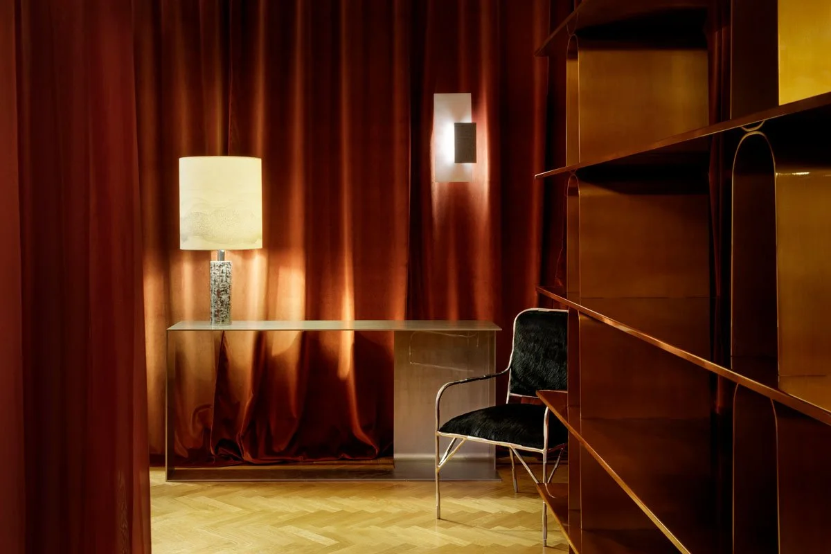 A cozy room with deep red curtains, a tall table with a lamp, a black velvet chair, and a wooden shelving unit.