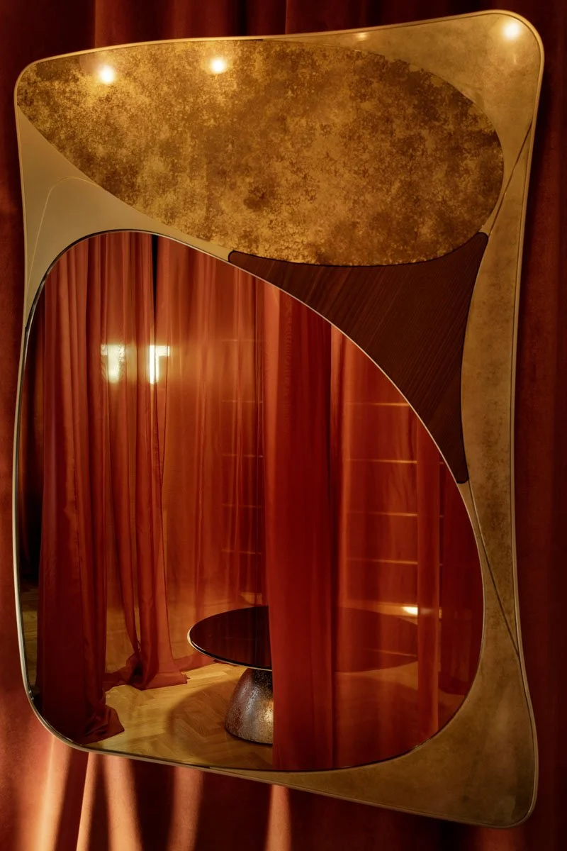 A decorative wall mirror with a gold and brown abstract design, reflecting a room with red curtains and a black round table.