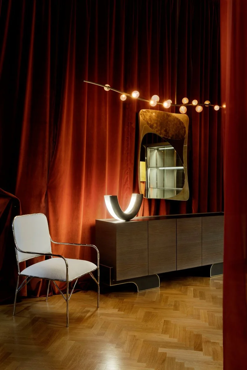 Interior of a room with red curtains, a mid-century modern white chair with metallic arms, a dark wood sideboard with a curved base, a semi-circular illuminated sculpture, a large irregularly shaped mirror with a metallic frame, and a string of globe lights hanging above.