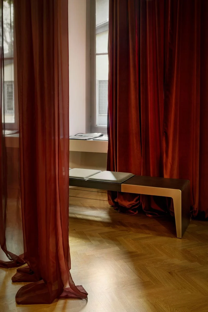 A room with red curtains, a wooden floor, a window, a bench with a cushion, and a small table with a magazine on it.
