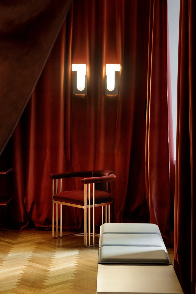A dimly lit room with heavy red curtains, a modern burgundy chair with gold metal frame, and a white lounge chair in front of the curtains.