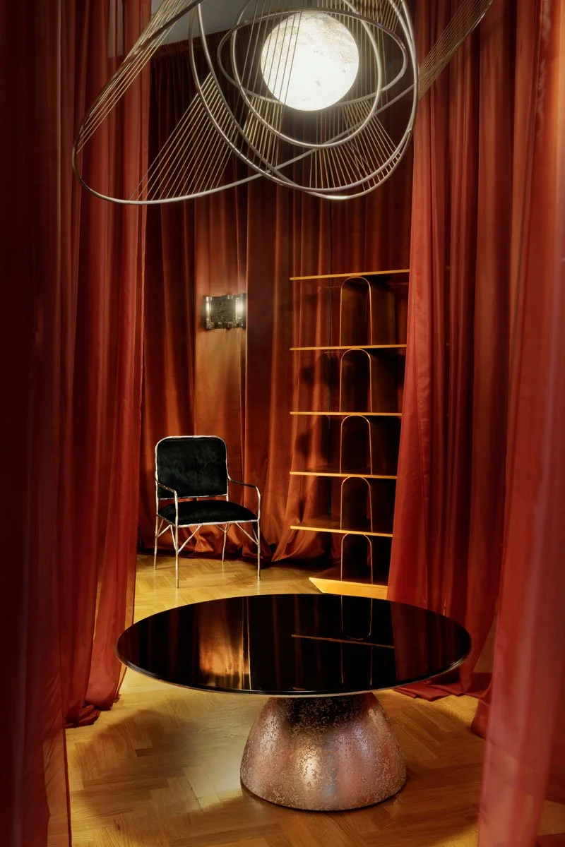 An interior room with red curtains, a black chair with a metal frame, a round black table with a textured base, a gold bookshelf, and a modern ceiling light fixture.