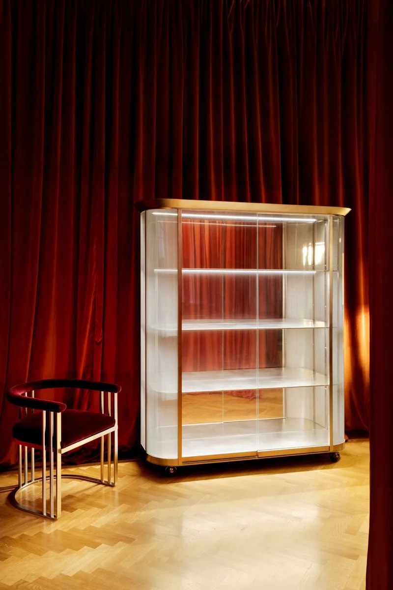 Empty glass display cabinet on wheels with wooden trim, beside a modern black and gold chair, in front of closed red curtains on a wooden floor.