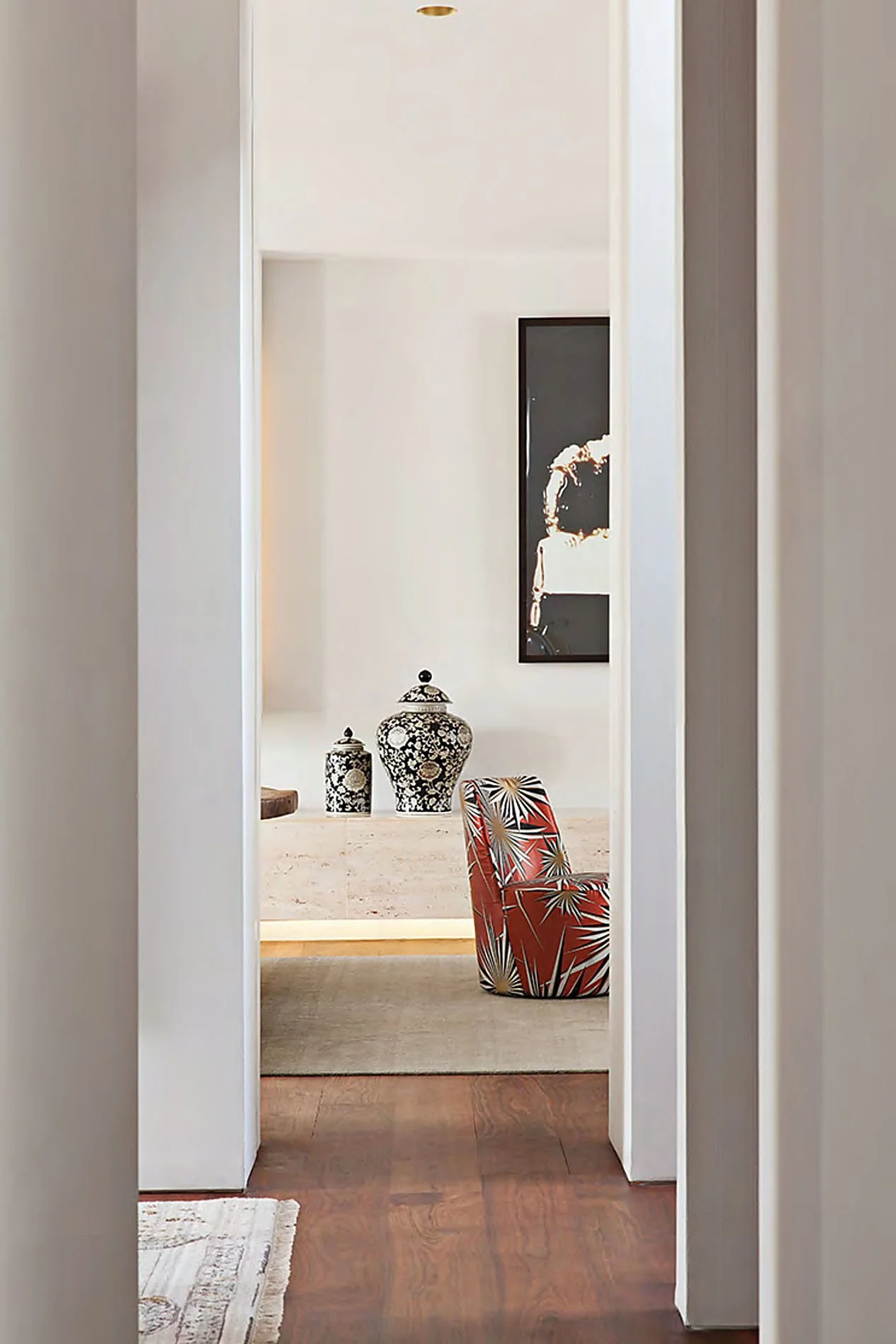 A hallway view leading to a living area with a patterned chair and decorative vases, framed art on the wall, and hardwood floors.