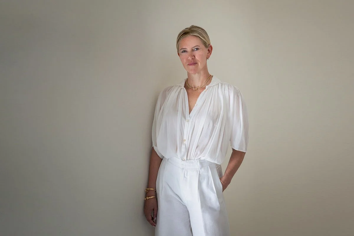 A woman with short blonde hair standing against a plain light-colored wall, wearing a white blouse and white high-waisted pants, looking directly at the camera with a slight smile.
