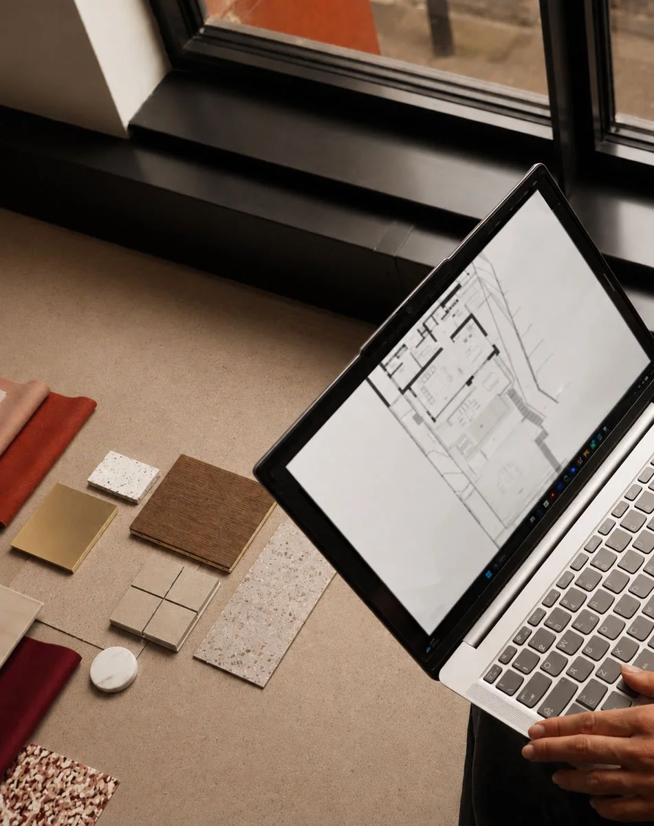 Person designing a building layout on a laptop with architectural drawings, fabric samples, and material samples on a beige surface near a window.