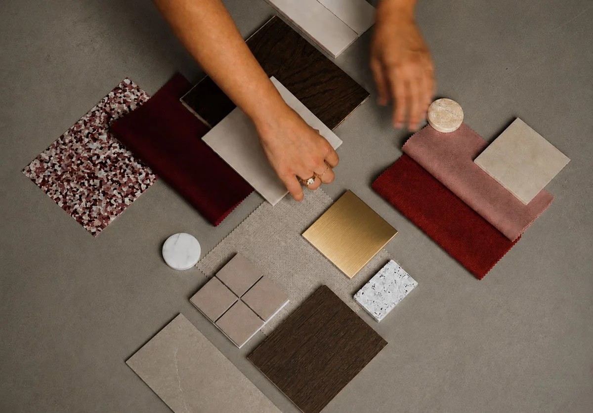 Hands arranging various small samples of fabric, tile, and metal on a gray surface.