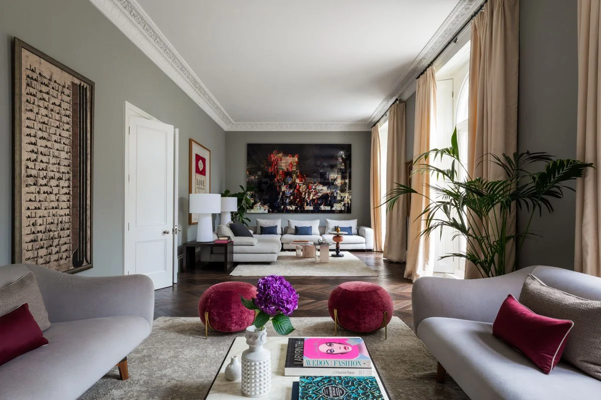 Spacious living room with light gray walls, large white sofa, two red velvet ottomans, two light gray sofas with red and beige pillows, a coffee table with magazines and a purple flower arrangement, large windows with cream curtains, a tall green plant, wall art including an abstract painting and a framed picture, and modern decor.