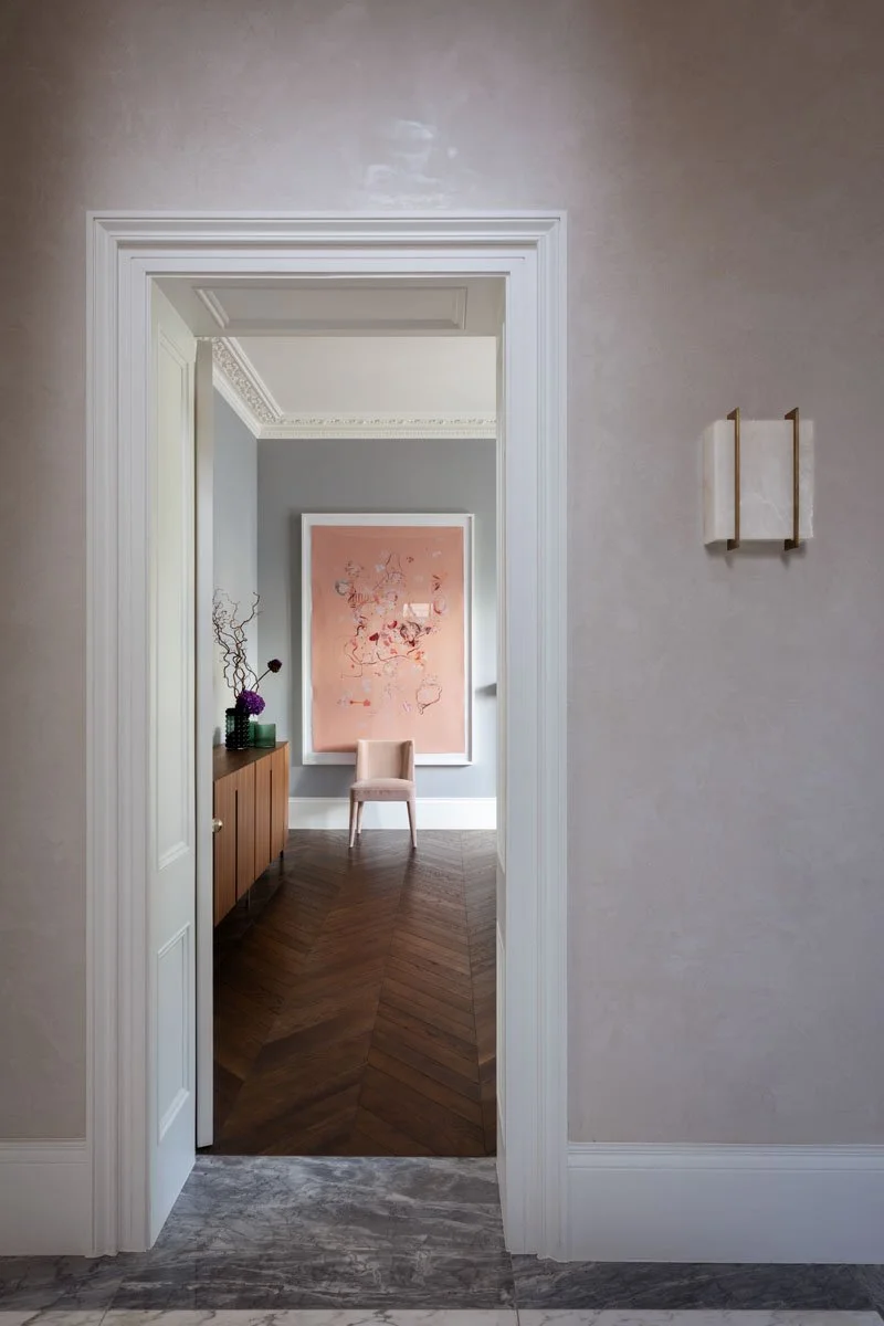 View through a doorway into a room with a modern interior, featuring a pink abstract painting on the wall, a beige chair, and a wooden sideboard with decorative items and flowers.