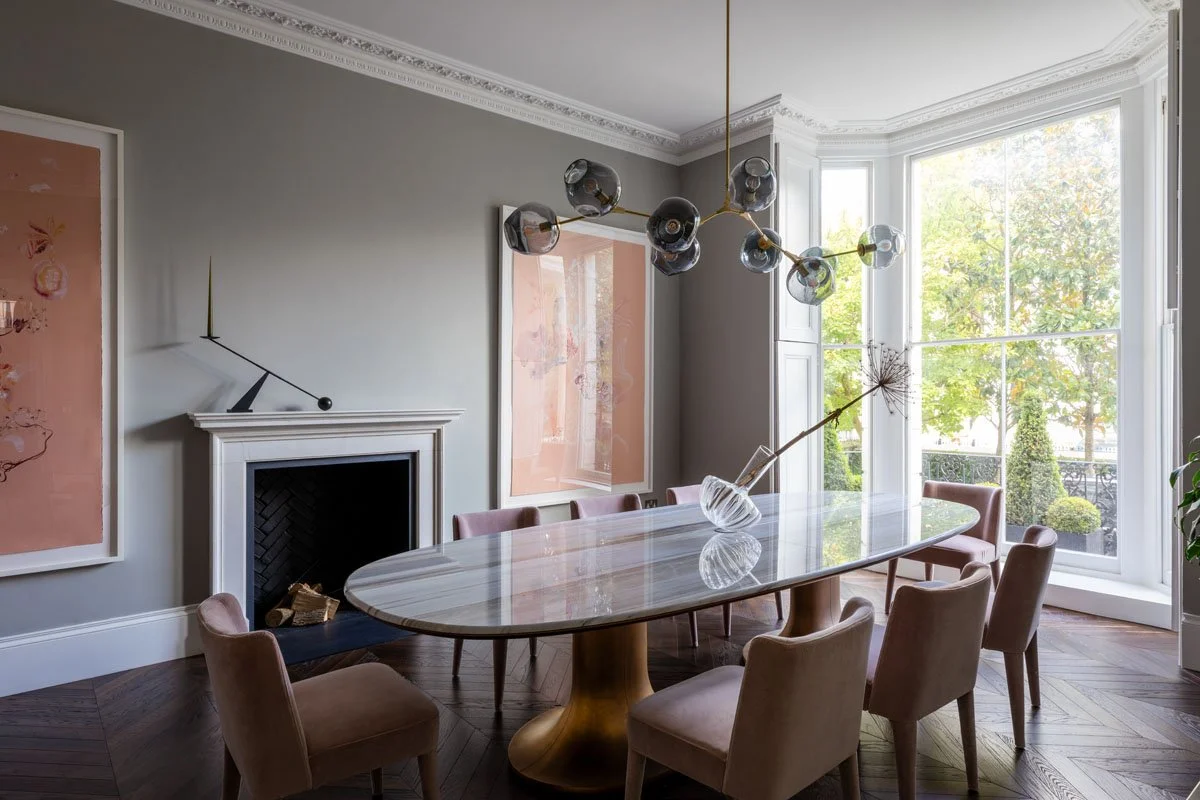 A modern dining room with a marble oval table, surrounded by beige upholstered chairs, featuring a glass pendant light fixture, large bay window with greenery outside, and artwork on the walls.