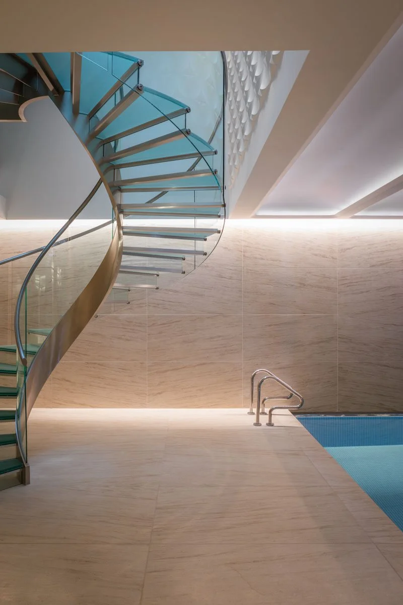 Modern indoor swimming pool with a curved glass spiral staircase and beige tiled walls and floor.