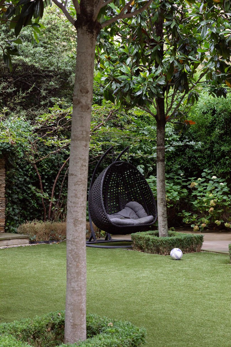 A backyard with a green lawn, two trees, a hanging egg-shaped chair with a cushion, and a soccer ball on the grass.
