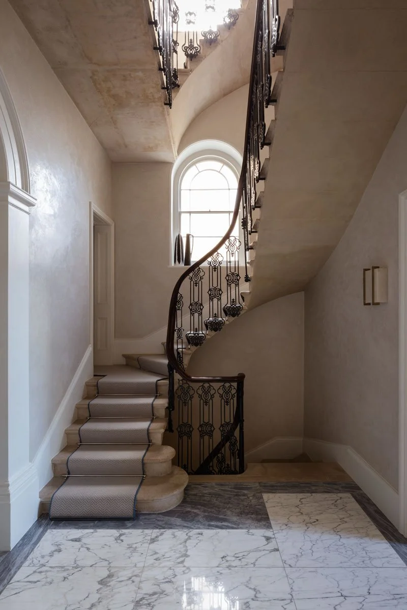 Interior view of a staircase with a curved, wrought-iron balustrade, carpeted steps, a large arched window allowing natural light, and marble flooring in a luxurious home.