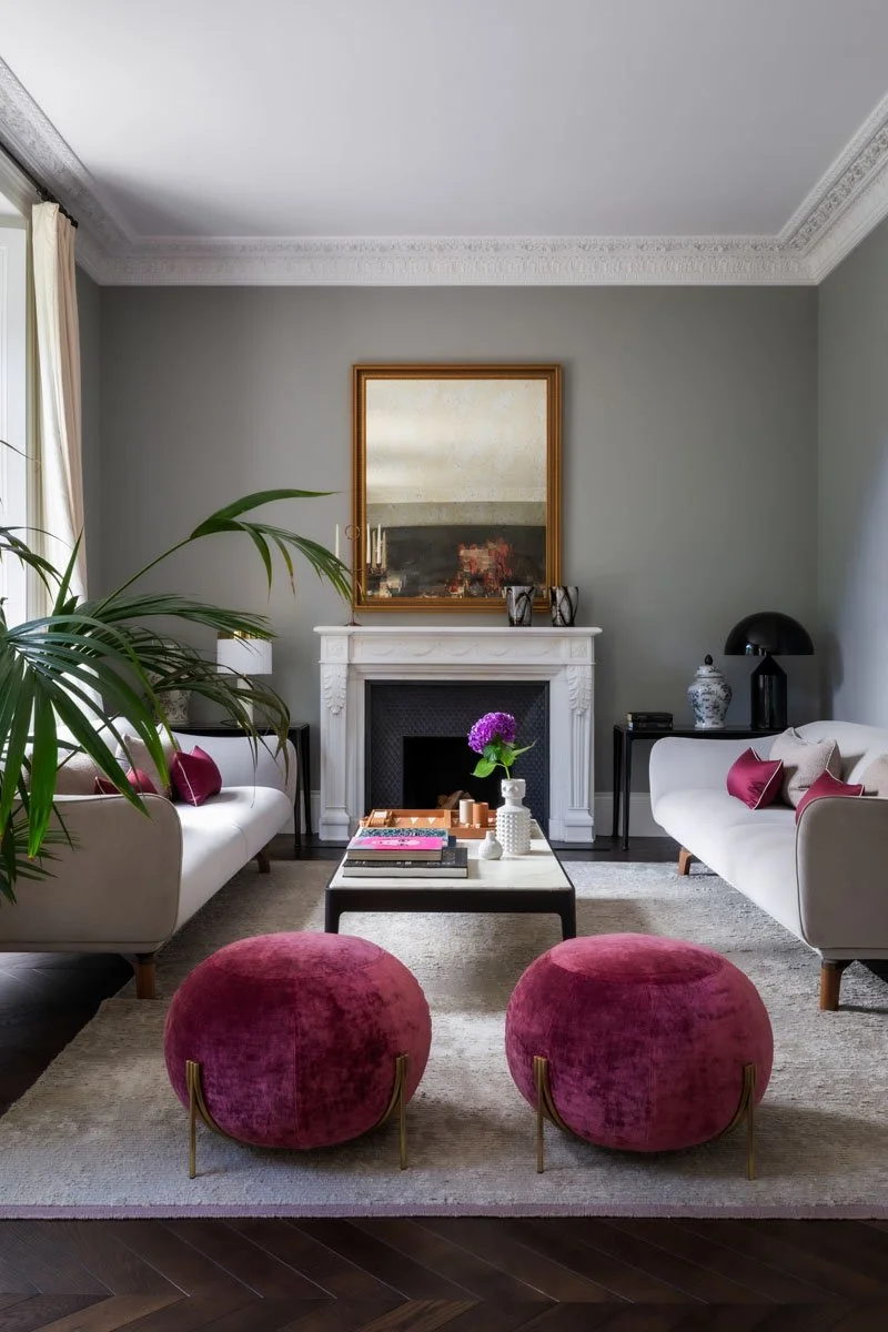 A living room with two white sofas, pink pillows, and a black coffee table with books and a purple flower. There are two pink velvet ottomans at the front, a fireplace with a mirror above it, and a green plant on the left. The room has crown molding and large windows.
