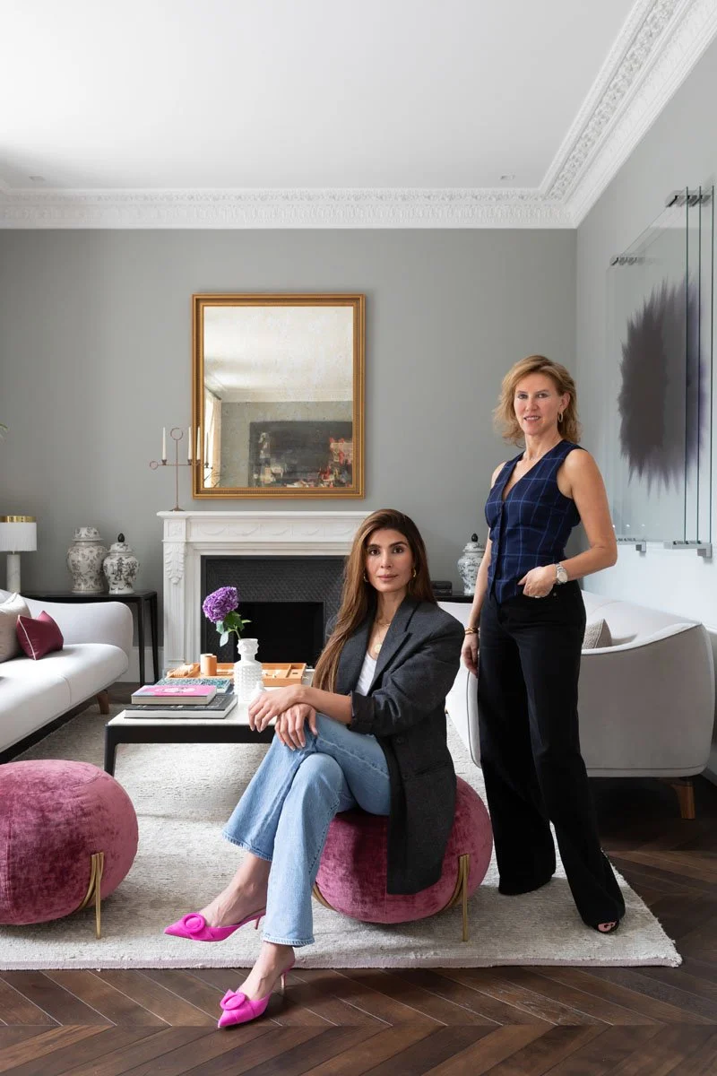 Two women in a well-decorated living room, one sitting on a pink ottoman with pink shoes, the other standing, both smiling. The room features modern furniture, a fireplace with a mirror above, and decorative vases.