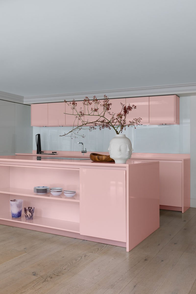 Modern kitchen with pink cabinets, a white vase with a face sculpture holding a branch, and open shelves with bowls and dishes, on a wooden floor.