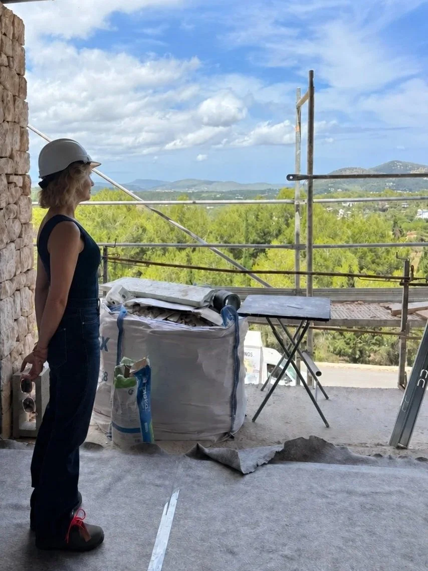 A woman in construction gear, including a white safety helmet, stands on a building site with a scenic view of greenery and mountains in the background. She is holding sunglasses and looking towards the landscape, near scaffolding and construction materials.