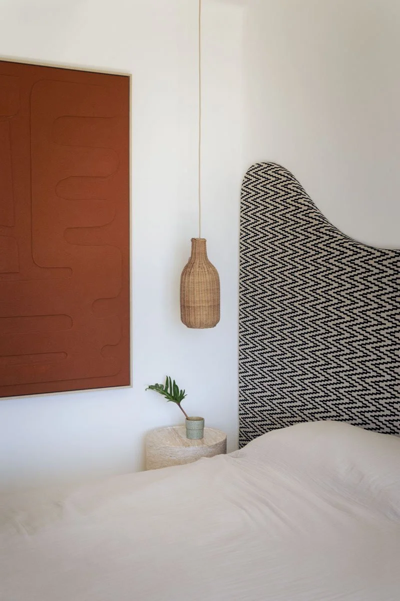 A bedroom wall with a framed brown artwork, a hanging wicker lamp, a small plant in a cup, and a bed with a patterned headboard.