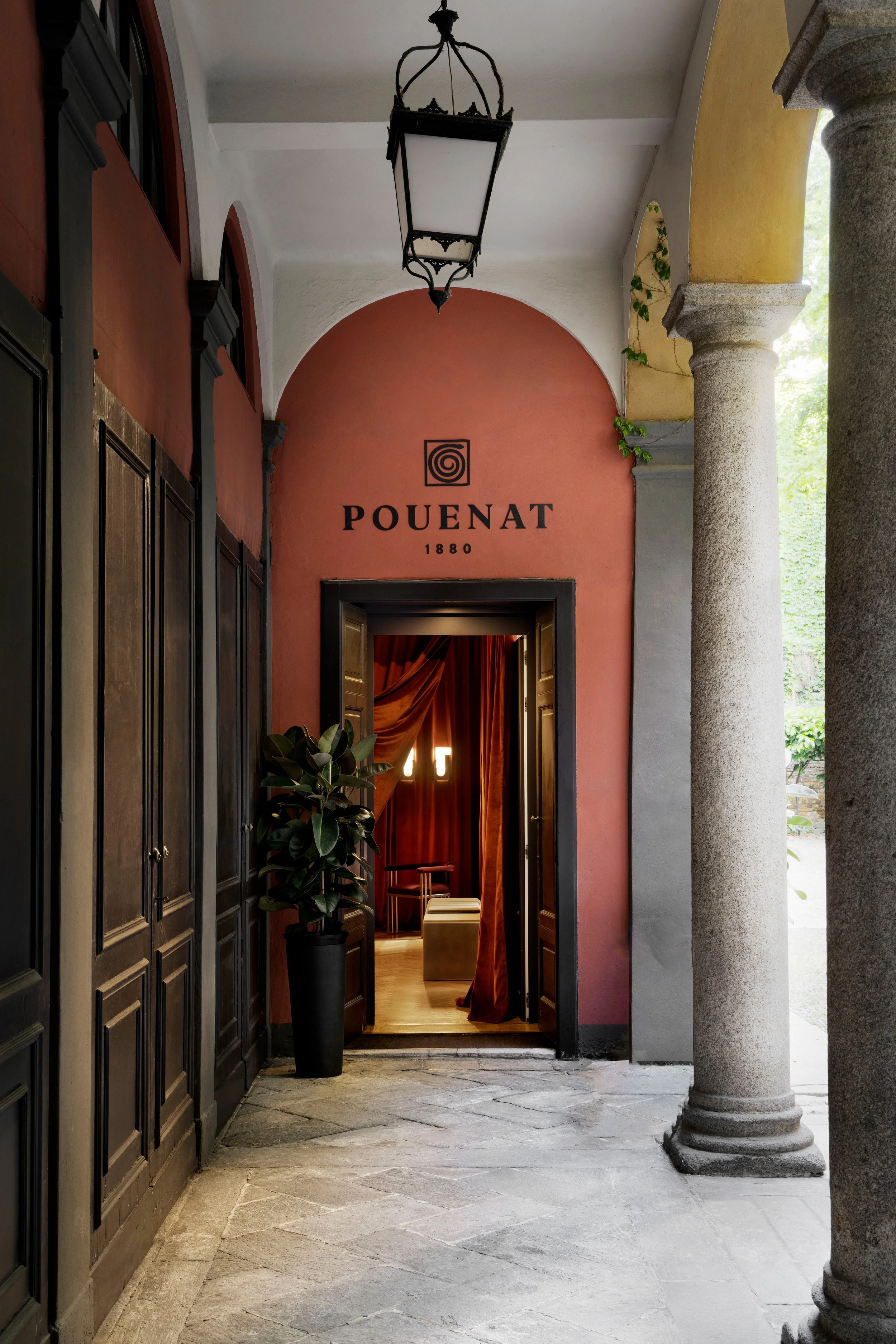 A corridor with stone flooring, dark wooden cabinets on the left, and stone columns on the right. An arched doorway with red curtains leading to a room with warm lighting. A hanging lantern-style light fixture from the ceiling, and the wall above the door has the word 'POUENAT 1880' written with a spiral logo above.