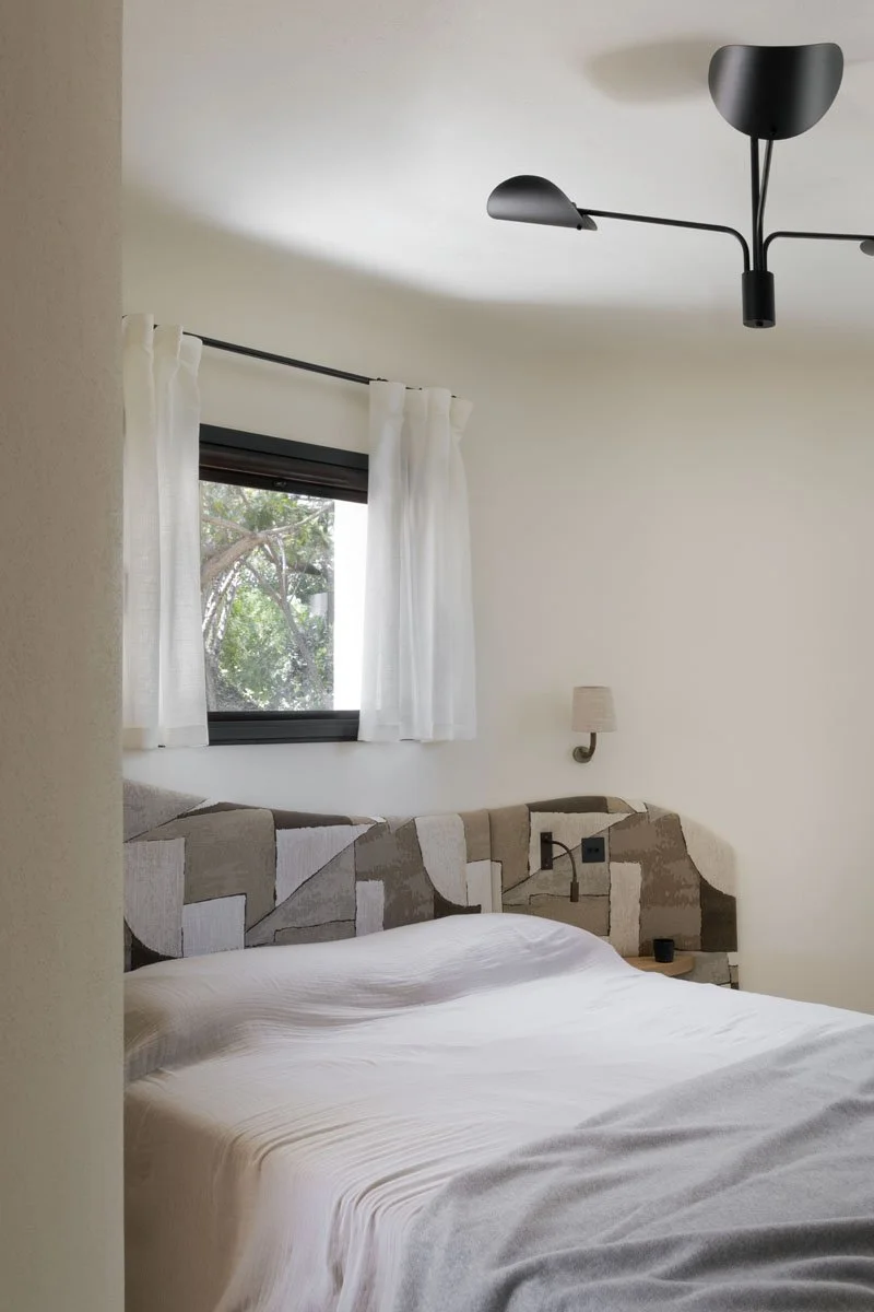 A bedroom with a window covered by white curtains, a beige and brown patterned headboard, a white bedspread, a wall-mounted lamp, and a black ceiling light fixture.