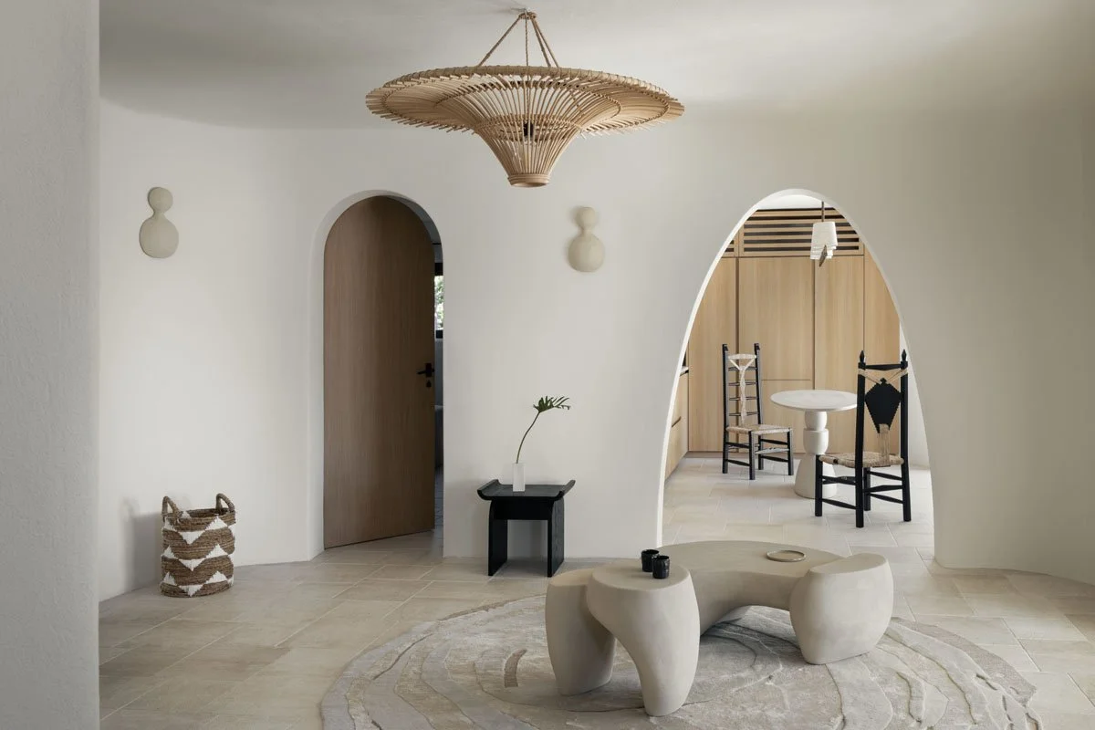 Minimalist living space with a curved white wall, a beige textured tiled floor, a modern sculptural beige coffee table on a neutral rug, a small black side table with a plant, woven basket, and a hanging wooden light fixture.