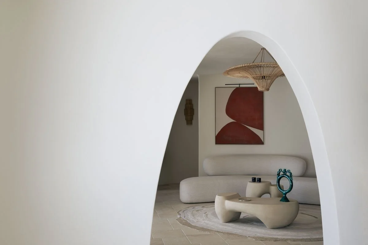 Modern living room with white curved sofa, abstract artwork, and sculptural coffee table seen through an arched doorway.