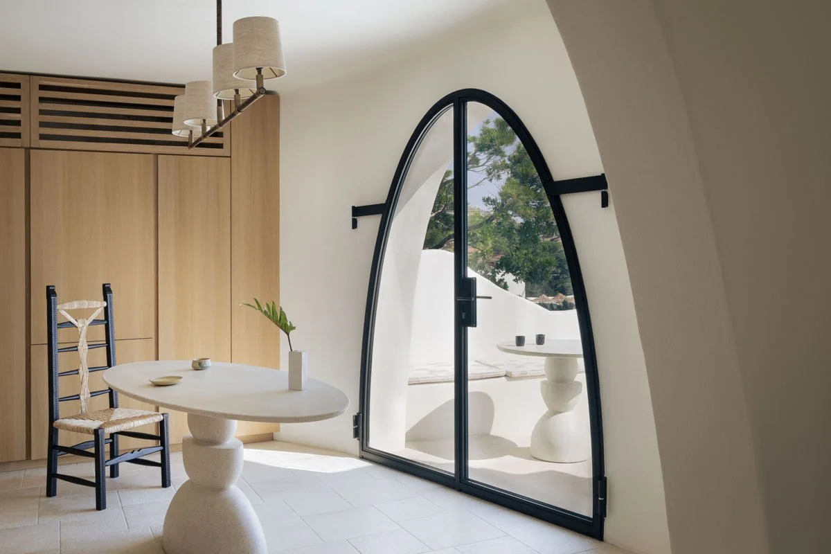 Minimalist room with a round white table, a black chair with a woven seat, a wooden closet, a modern chandelier, and a large arched glass door leading to an outdoor space with trees.