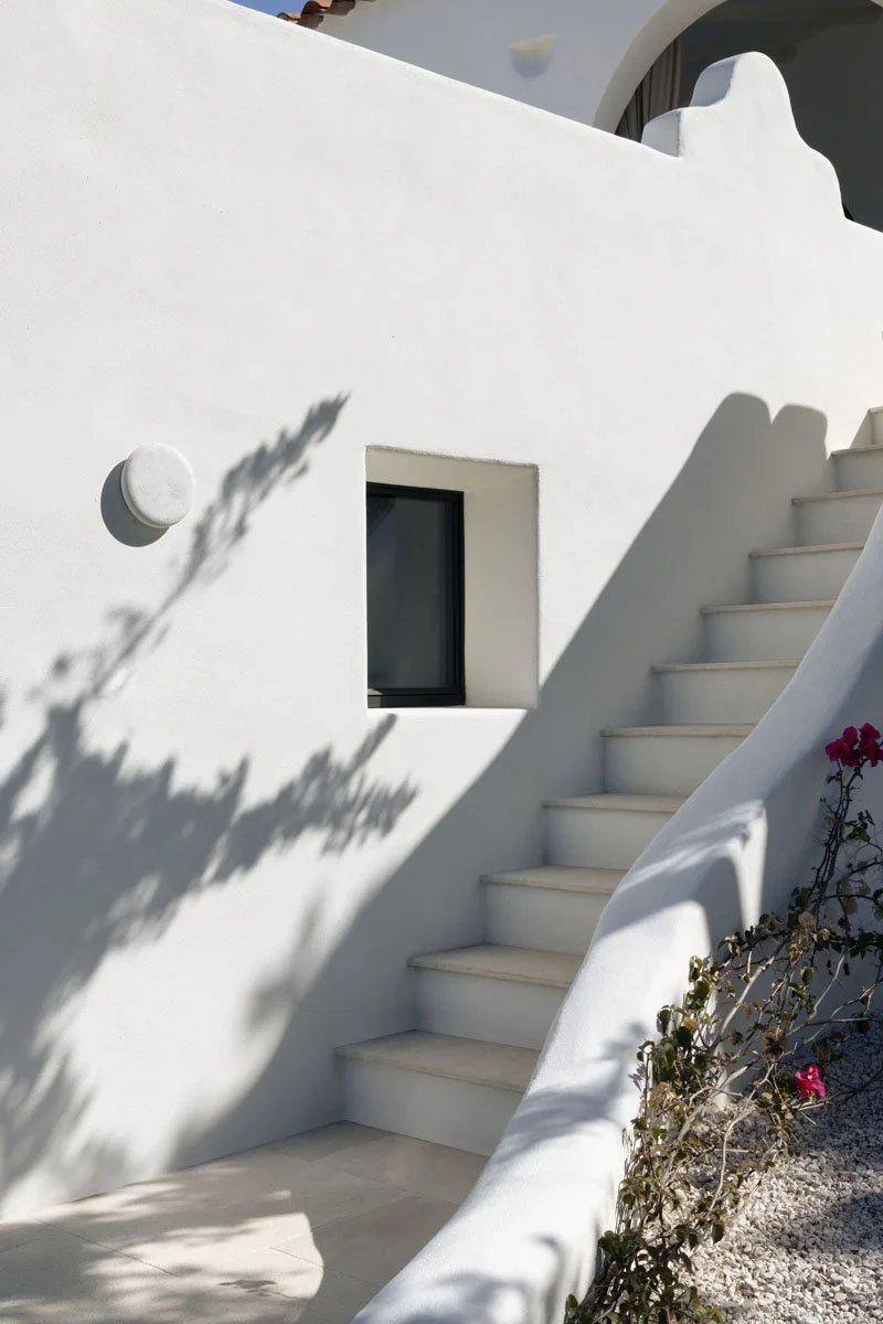 White stucco building with a staircase curving along its side, a small window, and a round wall-mounted light fixture. Shadows of plants are cast on the wall, with some pink flowers visible near the stairs.