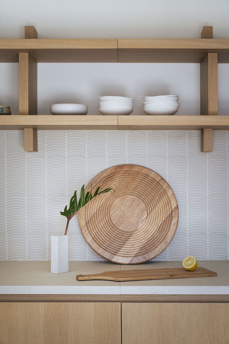 A minimalist kitchen shelf display featuring white bowls stacked on the middle shelf, a potted green plant, a lemon half, and a wooden cutting board against a patterned wall.
