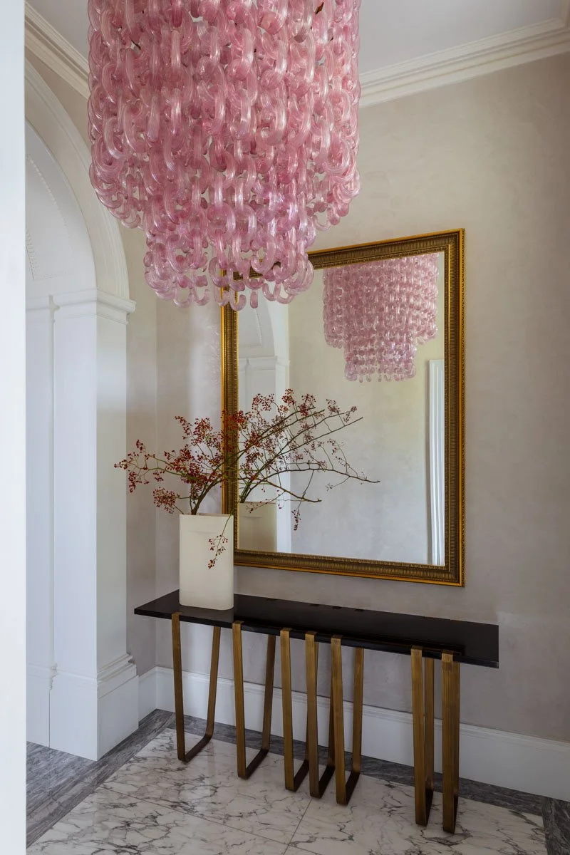 A pink chandelier hangs from the ceiling, reflected in a large gold-framed mirror on the wall. Below the mirror, a black console table with gold legs holds a white vase with red berries and branches.