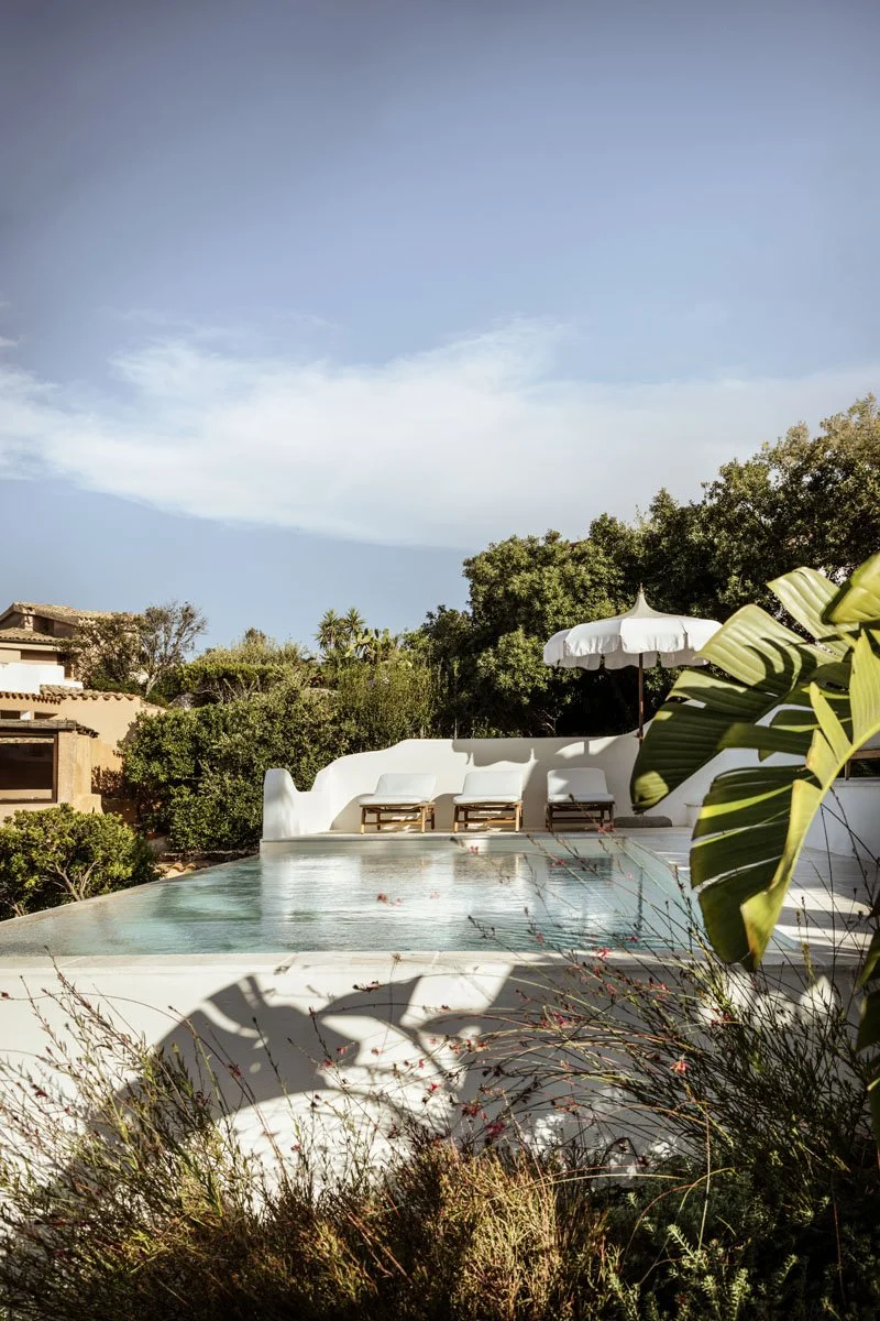 A backyard with an infinity pool, surrounded by greenery and a white curved wall, with lounge chairs and an umbrella. The sky is partly cloudy with a blue hue.