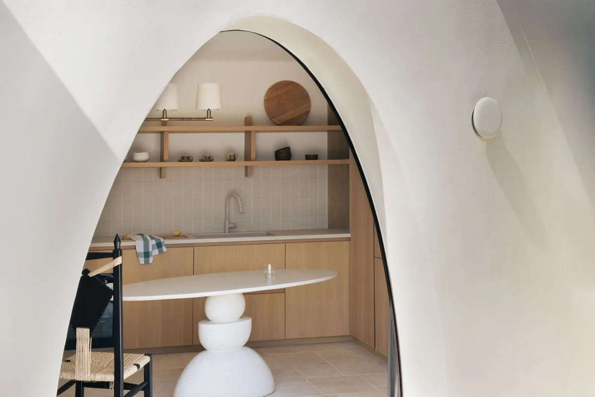 Indoor kitchen seen through an arched opening, with wooden cabinets, open shelving, a small sink, and a white table with rounded base.