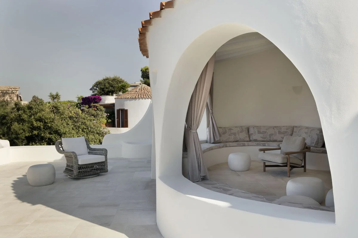 Outdoor terrace with white curved walls, cushioned seating, a wicker armchair, and small white stools, overlooking greenery and neighboring buildings under a clear sky.