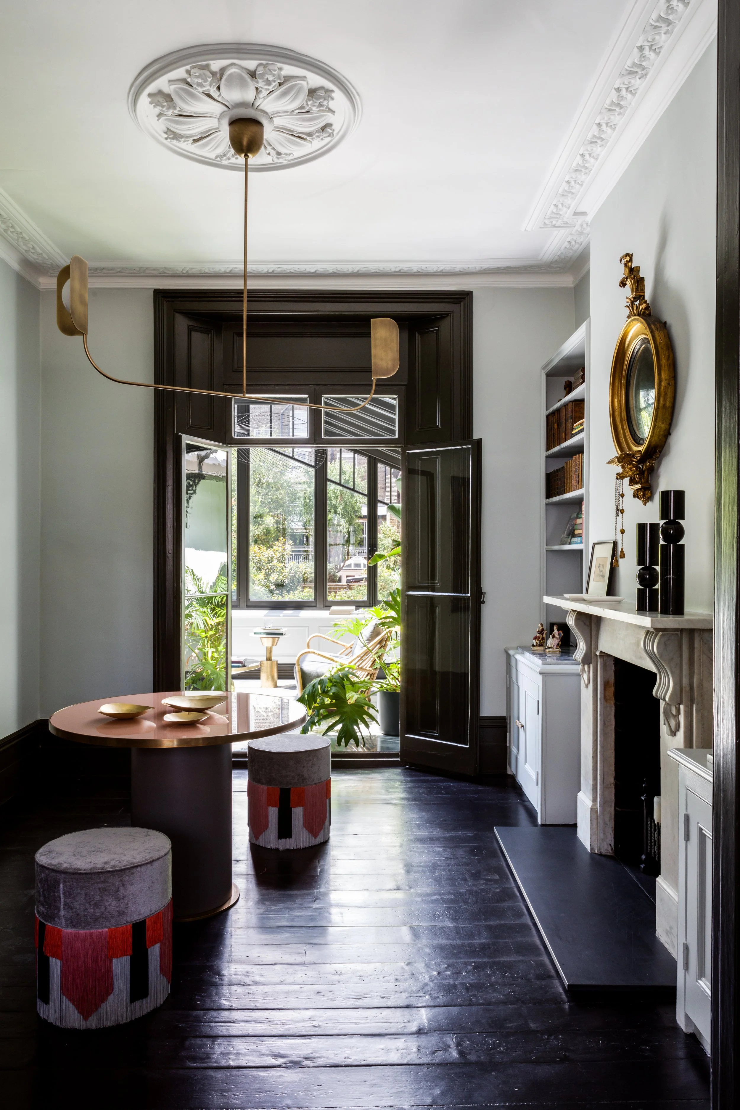 A living room with a round pink table and two cylindrical stools with a geometric red, black, and white pattern. The room has dark wooden floors, a white mantel with decorative items, a gold-framed mirror, and a large window with black trim opening to a patio with garden plants.